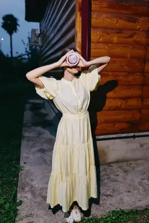 Уличный портрет женщины в светло-желтом платье с фотоаппаратом, позирующей у деревянной конструкции в сумерках для мрачного кинематографичного снимка