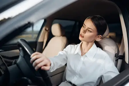 Женщина за рулем автомобиля, одетая в белую рубашку, сидит на водительском сиденье, положив руку на руль и закрыв глаза, что наводит на мысли об усталости или глубокой задумчивости
