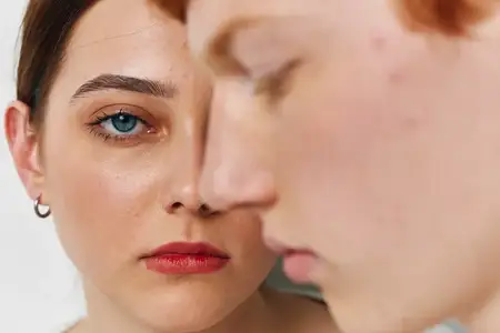 Портрет молодой женщины и мужчины с гладкой кожей, сосредоточенным взглядом и естественным макияжем, демонстрирующий крупные черты лица и эмоциональную напряженность на белом фоне