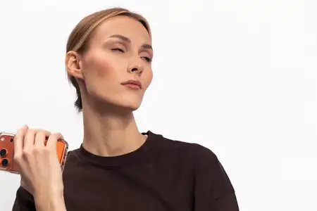 Спокойная молодая женщина в коричневом свитере держит в руке смартфон и отводит взгляд с закрытыми глазами, демонстрируя задумчивое выражение лица в простой студийной обстановке на белом фоне