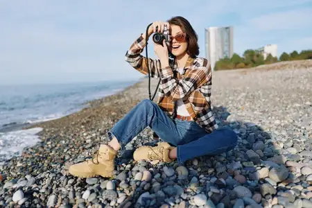 Сцена уличной фотографии: женщина с камерой на галечном пляже, наслаждающаяся морскими видами и расслабленным образом жизни при дневном свете с живописной береговой линией и горизонтом