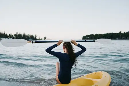 Женщина в темном гидрокостюме сидит на желтом каяке лицом к воде, поднимая весло над головой; вокруг спокойная вода и безмятежный природный пейзаж