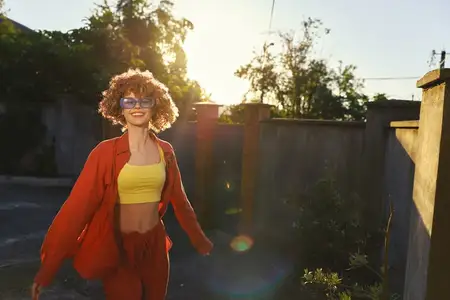 Она на улице в солнечный день, в жёлтом топе и оранжевой куртке, мягко улыбается, прогуливаясь по залитой солнцем дорожке, создавая яркий жизненный момент
