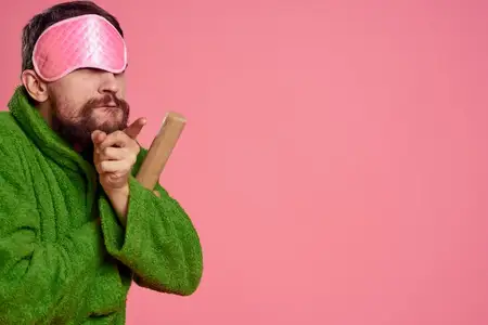 Мужчина в розовой маске для сна на лице, в зелёном халате, со скалкой в руке