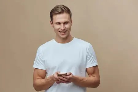 Улыбающийся мужчина держит смартфон, одет в белую футболку, смотрит в сторону на бежевом фоне, повседневный вид, концепция коммуникационных технологий, образ жизни цифрового кочевника, удаленная…