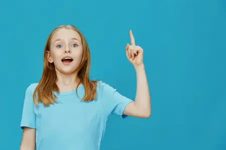 Веселая юная девушка в голубой футболке проявляет энтузиазм, подняв палец, на фоне ярко-голубого фона