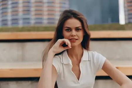Молодая женщина в белом поло улыбается на улице на фоне города, при естественном свете, в летнем стиле, повседневный модный портрет, передающий уверенное и расслабленное настроение
