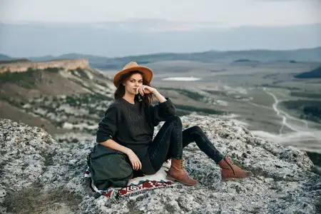 Женщина с рюкзаком и в шляпе сидит на краю горной скалы, наслаждаясь живописным видом