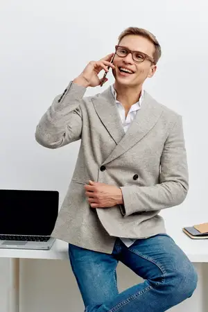 Улыбающийся мужчина разговаривает по смартфону, одетый в повседневный серый пиджак и очки, сидит рядом с ноутбуком в современном офисе, демонстрируя расслабленную позу и позитивное настроение