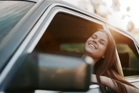 Радостная молодая женщина с длинными волосами, погруженная в момент во время поездки, улыбается, высунувшись из окна автомобиля в мягком свете заката, подчеркивая свободу и приключение