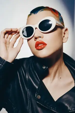 Крупный план женщины с яркими разноцветными волосами, белыми солнцезащитными очками, глянцевыми красными губами и черной кожаной курткой на чистом фоне, демонстрирующий смелый современный стиль