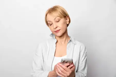Пожилая женщина держит смартфон в помещении, сосредоточена на текстовом общении, лёгкая улыбка, расслабленные руки, мягкий студийный свет, чистый нейтральный фон