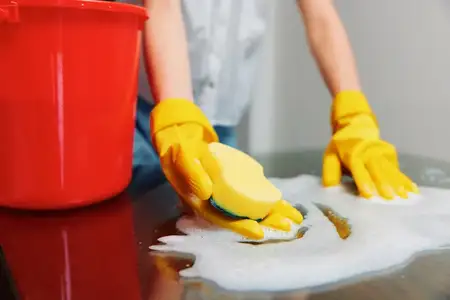 Уборщица в жёлтых перчатках моет стол с мылом и ведром мыльной воды перед красным ведром