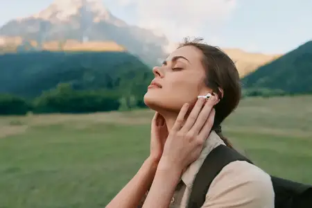 Одинокая молодая женщина стоит на широком зеленом поле на фоне гор, в наушниках и с закрытыми глазами, погружаясь в покой и осознанность в ярком природном окружении