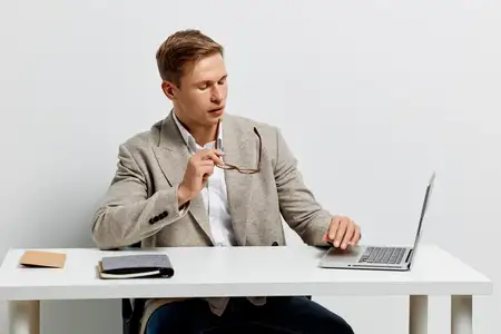 Молодой человек работает за ноутбуком в повседневной куртке, сидит за белым столом, изолирован на белом фоне, сосредоточен