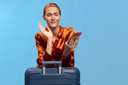 Молодая женщина в оранжевой рубашке выражает разочарование, держа телефон рядом с синим чемоданом на синем фоне