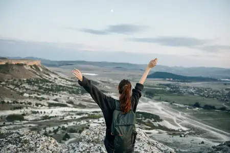 Женщина, раскинув руки в стороны, обнимает бескрайнюю горную долину внизу, застыв в восхищении от захватывающего вида