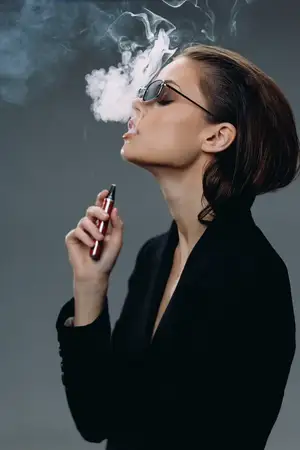 Стильная женщина в солнцезащитных очках парит, выдыхая густой дым в минималистичном сером окружении, что передает современный образ жизни и уверенность
