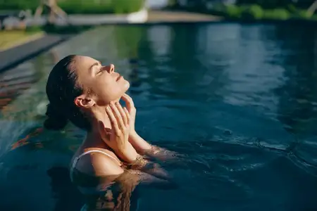 Сцена у открытого бассейна с женщиной в воде, солнечный свет на коже, расслабленное выражение лица, наслаждение отдыхом, атмосфера на открытом воздухе, естественные тона, пышная зелень…