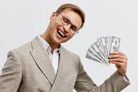 Веселый мужчина в очках и светлом пиджаке держит веер из долларовых купюр, широко улыбается на нейтральном фоне