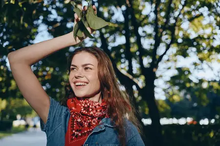 Женщина в стиле лайфстайл, одетая в современную богемно-вестерн джинсовую уличную одежду с красной банданой, ярко улыбается на открытом воздухе под пятнистым солнечным светом, передавая беззаботное…