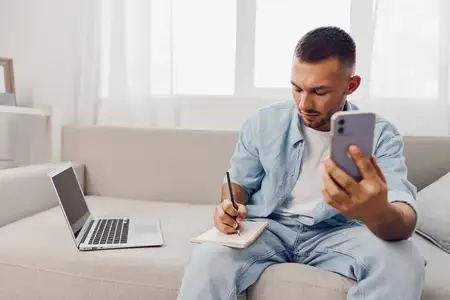 Целеустремленный мужчина в повседневной одежде использует смартфон и блокнот для повышения продуктивности в стильном домашнем офисе