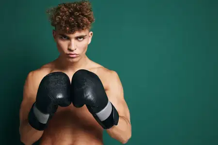 Спортивный молодой человек без рубашки с вьющимися волосами позирует с черными боксерскими перчатками в защитной стойке