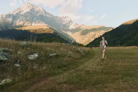 Женщина-туристка прогуливается по лугу в сторону суровых гор, с рюкзаком наготове, в легкой outdoor-экипировке, сцена залита солнечным светом, вызывая ощущение здоровья, приключений и единения с…