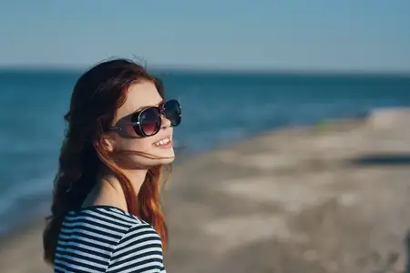 Женщина в очках и полосатой футболке стоит на балконе на закате, любуясь видом на море