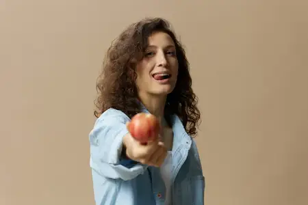 Милая, веселая, счастливая, кудрявая, красивая женщина в джинсах и повседневной рубашке протягивает яблоко к камере, позирует изолированно на бежевом пастельном фоне