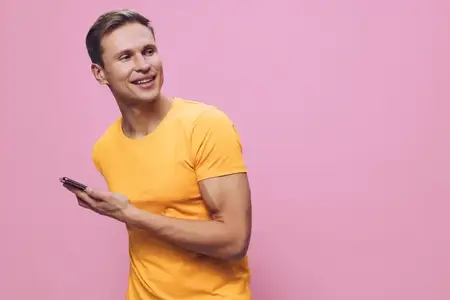Улыбающийся мужчина использует смартфон в желтой футболке, смотрит в сторону, стоит на розовом фоне, держит мобильное устройство, концепция цифрового кочевника с повседневным образом жизни и…