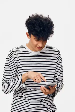 Молодой человек с вьющимися волосами, одетый в полосатую рубашку с длинным рукавом, сосредоточенно пользуется смартфоном на фоне однотонного белого фона, демонстрируя использование современных…