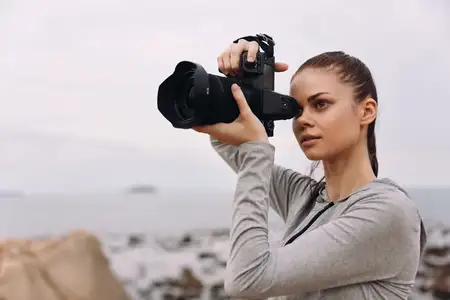 Молодая женщина в повседневной серой футболке с длинным рукавом фотографирует у океана, воплощая радость фотографии, любовь к природе и творчество в безмятежной обстановке
