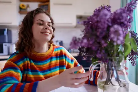 Яркая домашняя сцена: улыбающаяся женщина за обеденным столом, красочная одежда, мягкий естественный свет, цветы в вазе, расслабленное настроение, интимная сервировка завтрака, уютная домашняя…
