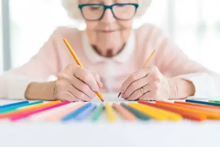 Пожилая женщина занимается арт-терапией в окружении ярких цветных карандашей