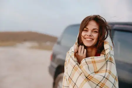 Женщина, укутанная в одеяло, улыбается перед машиной, путешествуя стильно и с комфортом в автомобильной поездке