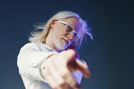 Пожилой мужчина с длинными седыми волосами и стильными очками игриво указывает на зрителя, одетый в безупречную белую рубашку