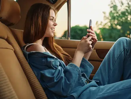 Радостная девушка в джинсовой одежде улыбается, пользуясь смартфоном в уютном автомобиле, передавая суть молодости, комфорта и современного образа жизни в солнечный день