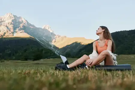 Безмятежная outdoor-сцена показывает молодую женщину, наслаждающуюся балансом между работой и жизнью: открытый ноутбук, телефон в стороне, на фоне зеленого поля и далеких вершин, что иллюстрирует…
