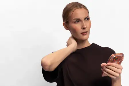 Растерянная молодая женщина со смартфоном в руке чешет шею