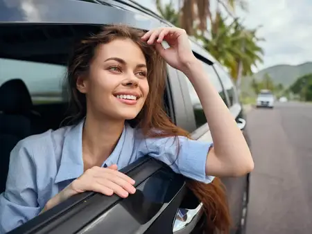 Улыбающаяся женщина, высунувшись из окна автомобиля, стоит на живописной дороге, вдоль которой выстроились пальмы