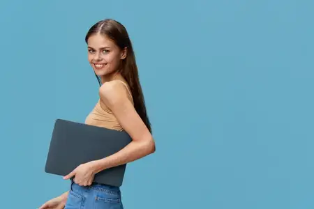 Улыбающаяся женщина в повседневном бежевом топе и синих джинсах держит ноутбук, глядя в камеру, позируя на фоне однотонного светло-голубого фона, который подчеркивает ее расслабленное и уверенное…