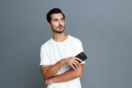 Задумчивый молодой человек держит смартфон в руке и смотрит вдаль, одет в простую белую футболку на минималистичном сером фоне