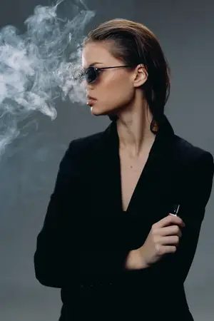Стильная женщина в солнцезащитных очках выдыхает дым в мрачной темной атмосфере на сером фоне, демонстрируя уверенность и загадочность