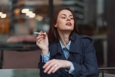 Повседневная уличная сцена: молодая женщина в джинсовой куртке отдыхает на террасе кафе, куря; городские огни размыты позади нее, создавая мрачную, урбанистическую атмосферу