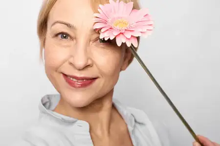 Женщина, красота, уход за кожей, цветок, крупный план, улыбка, лицо, здоровье, хорошее самочувствие, спа, зрелый портрет с розовым бутоном возле глаза