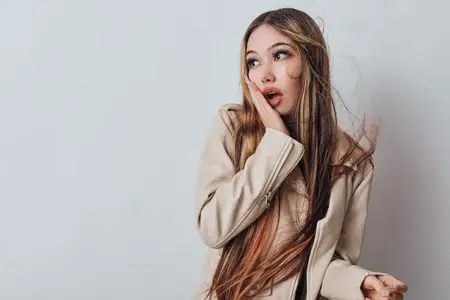 Молодая женщина в бежевой куртке с развевающимися волосами показывает удивление, приложив руку к лицу
