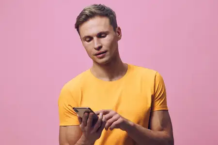 Молодой человек использует мобильный телефон в желтой футболке, сосредоточенно глядя на экран, держит устройство, разговаривает или переписывается