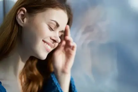Улыбающаяся женщина прячется с одеялом у окна, отдыхая в лучах утреннего солнца