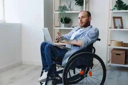 Мужчина в инвалидной коляске, фрилансер, улыбается, работая за ноутбуком дома
